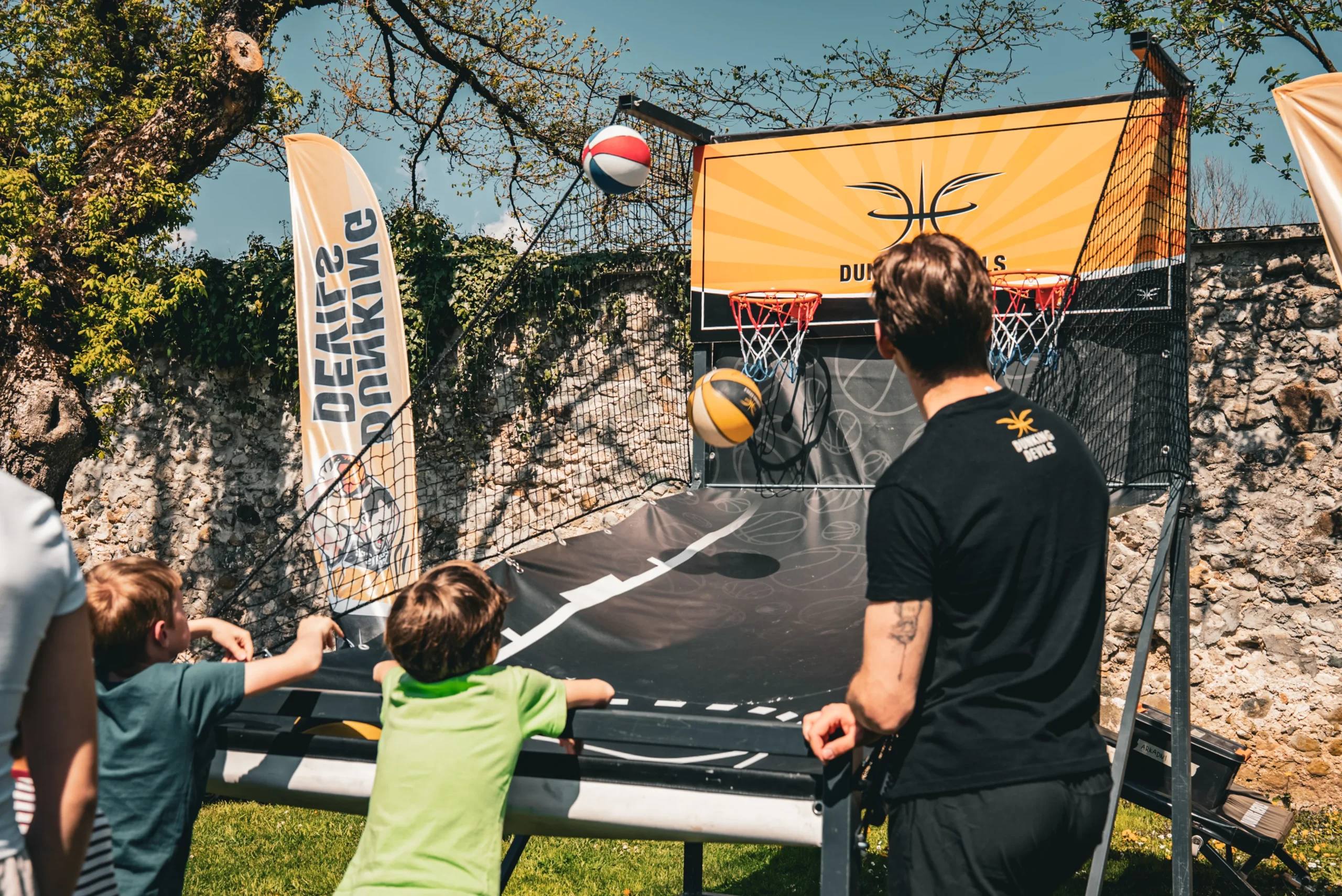 Children shoot at a Dunking Devils arcade hoop while a staff member helps.