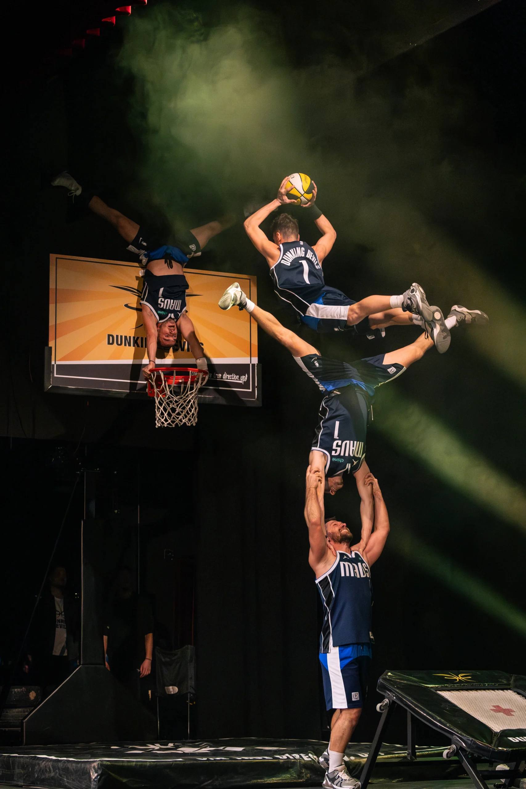 A Dunking Devils acrobat dunks over a teammate under green stage light.