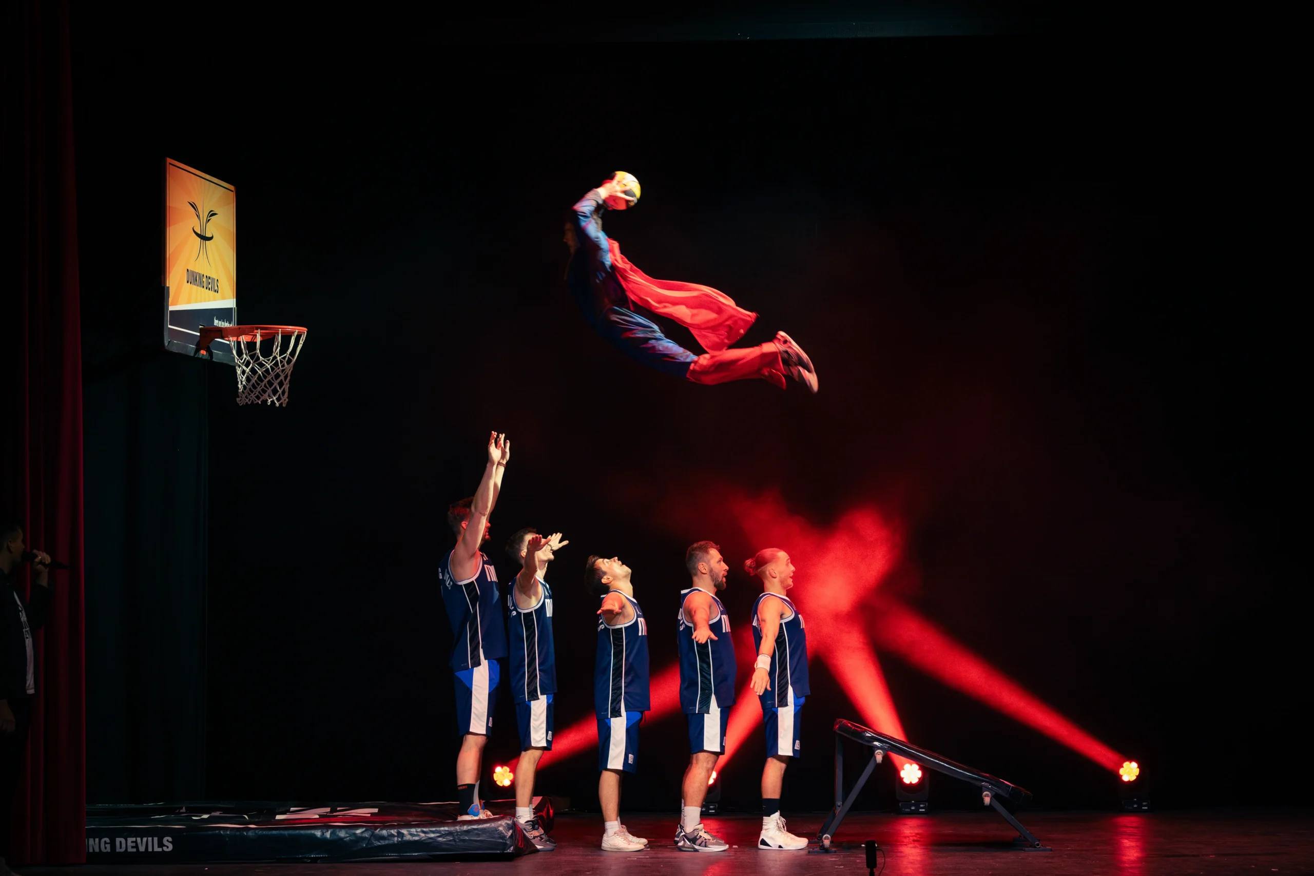 An acrobat flies towards the hoop above the cast under red stage lights.