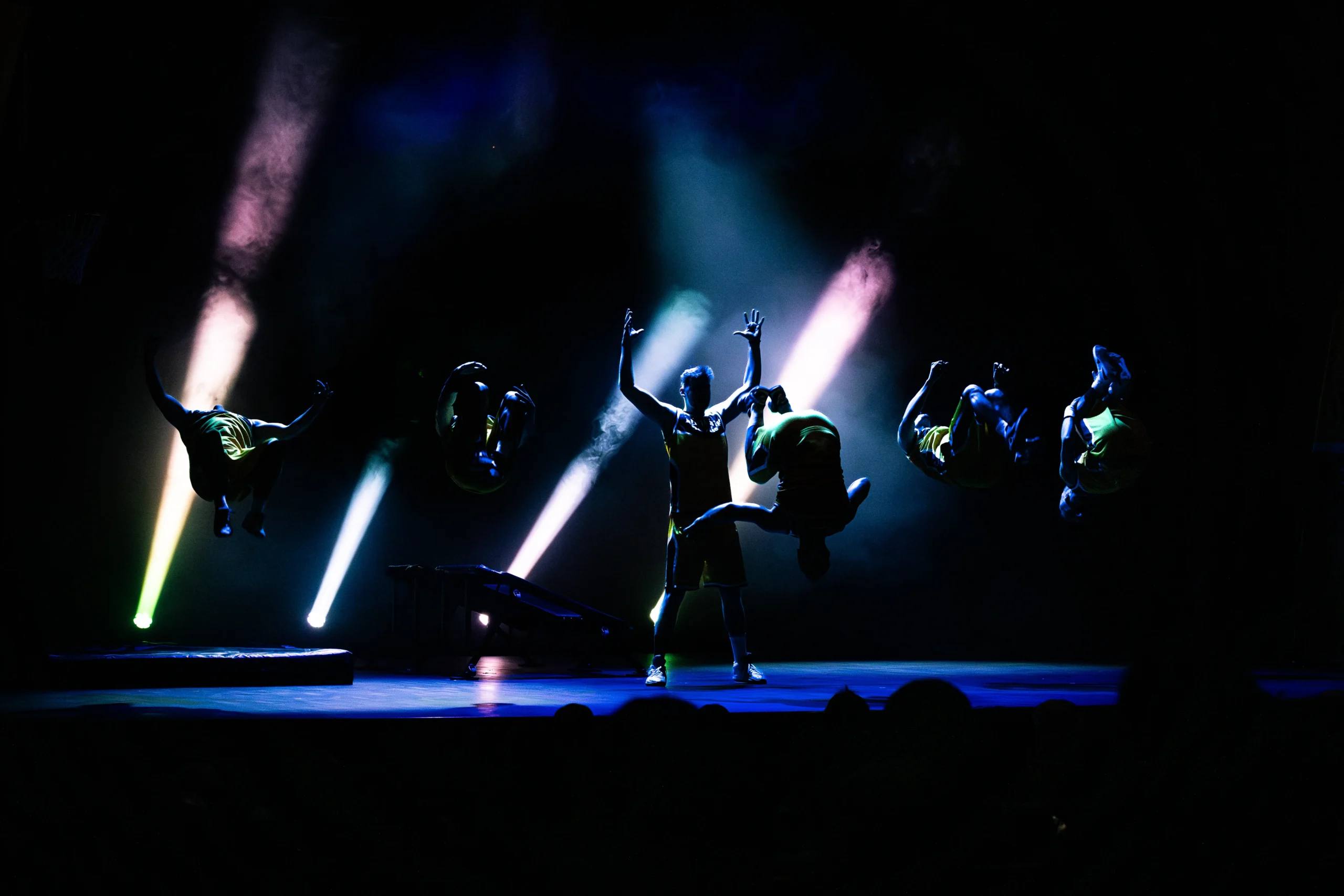 Performers frozen in silhouette between spotlight beams on the Špas Teater stage.