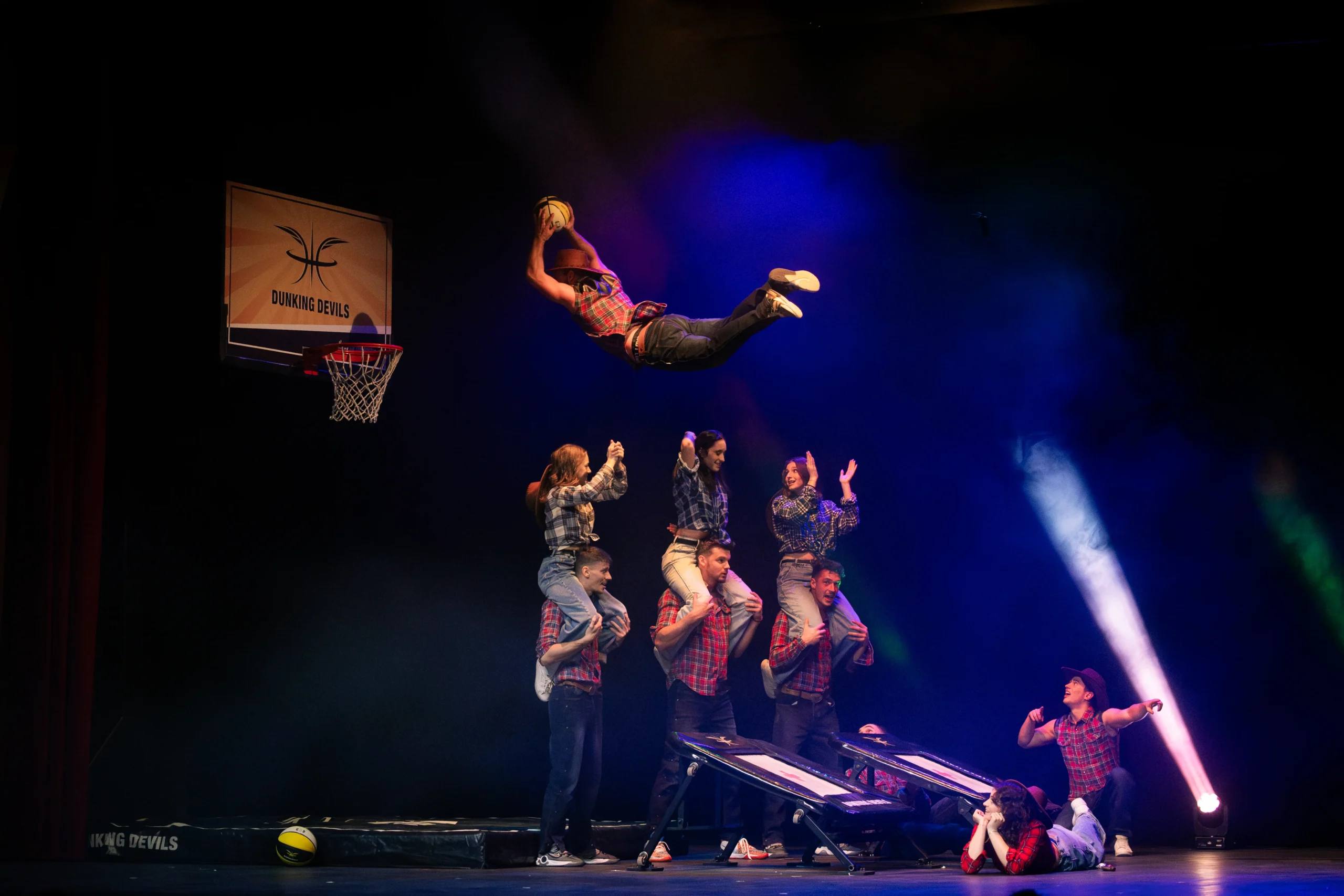 Dunking Devils acrobatic theatre show at Špas Teater: an acrobat flies above a human tower under blue stage lights, next to a basketball hoop.