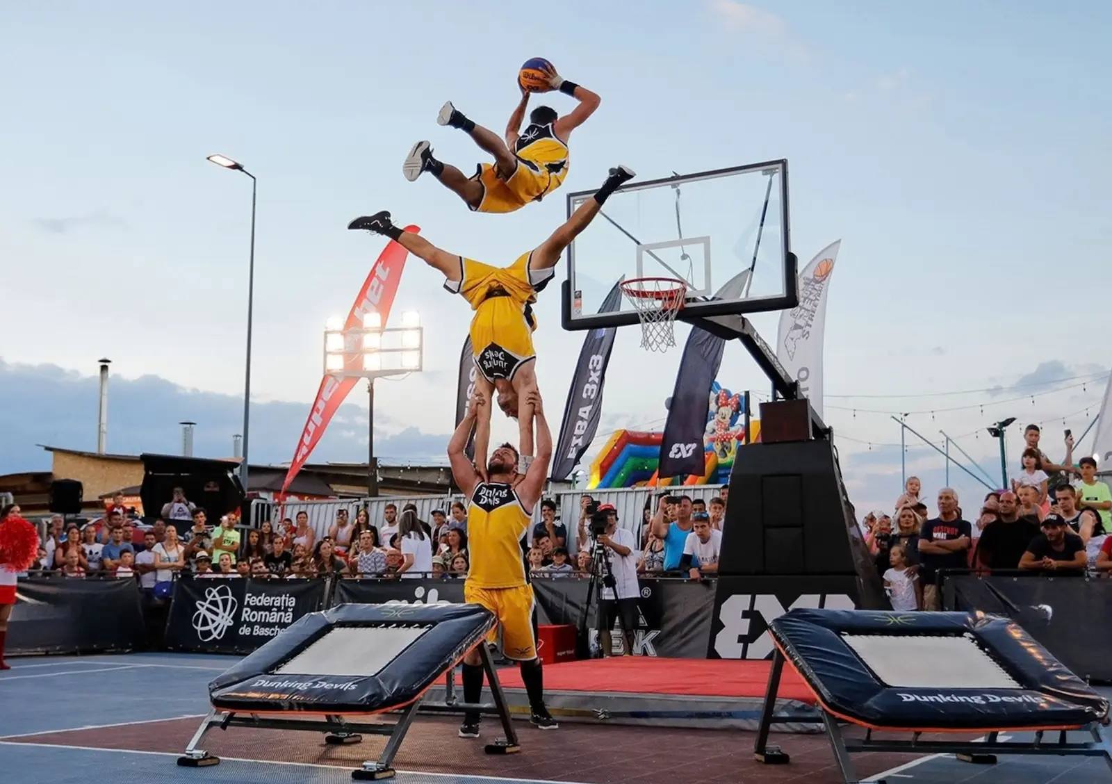 Dunking Devils acrobatic dunk show at a FIBA 3x3 event in Romania: one athlete flies over a human tower towards the hoop between two trampolines, with a crowd watching.