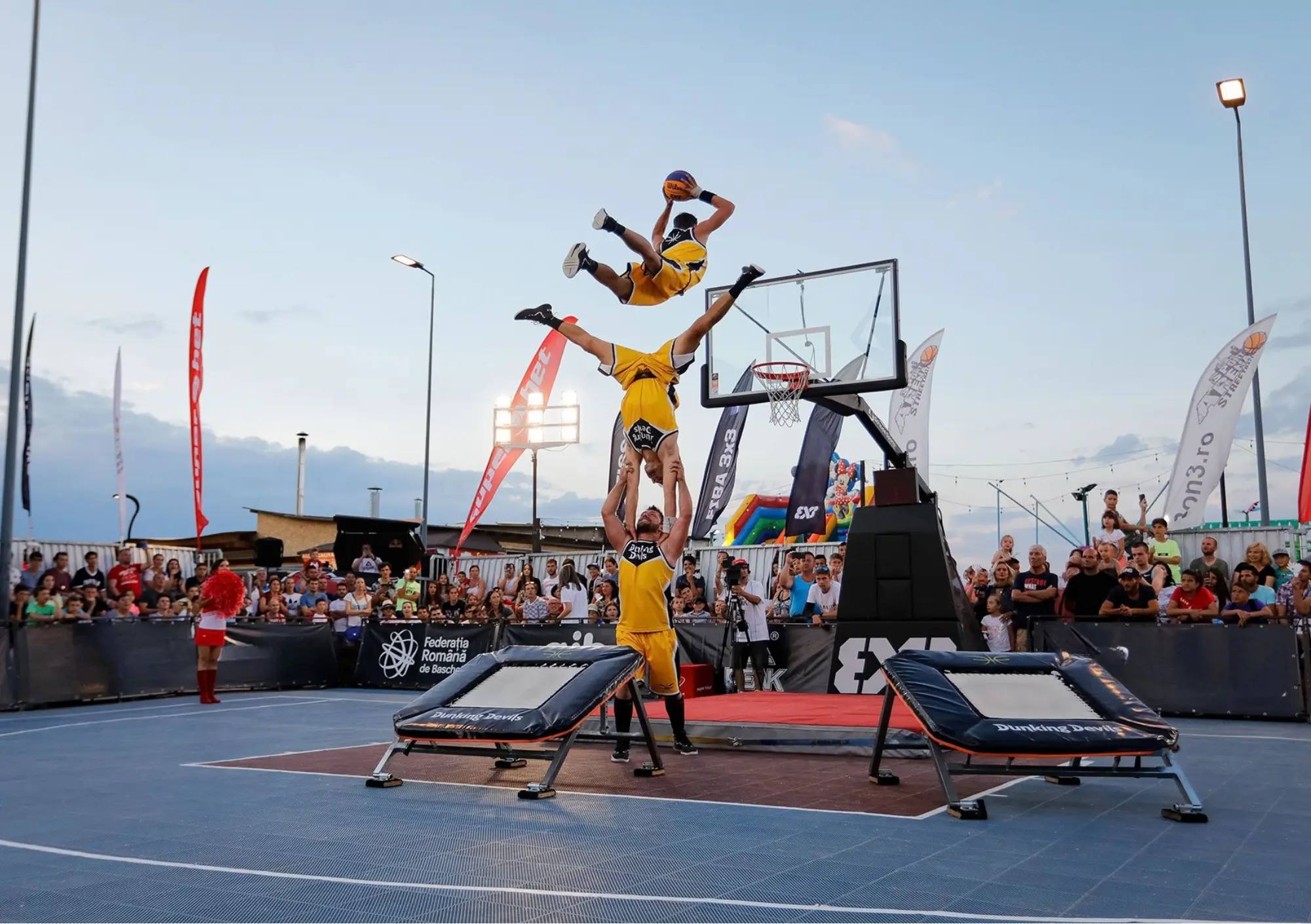 Dunking Devils acrobatic dunk show at a FIBA 3x3 event in Romania: one athlete flies over a human tower towards the hoop between two trampolines, with a crowd watching.