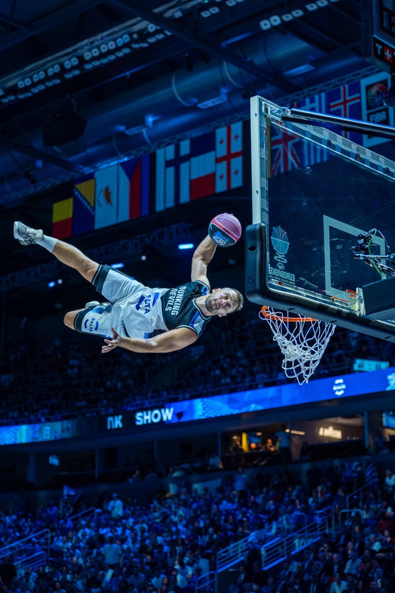 Dunking Devils athlete flying towards the hoop for a dunk during a EuroBasket 2025 halftime show, with the arena crowd below.