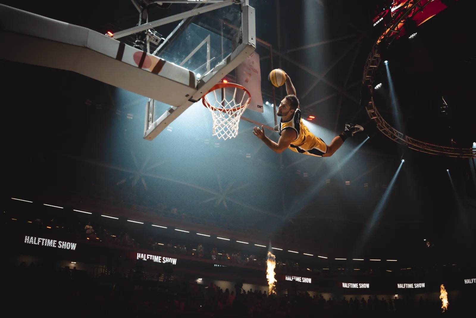 Ein Akrobat der Dunking Devils fliegt unter blauen Spots zum Ring, auf den Arenabanden steht „Halftime Show“, darunter Flammen.