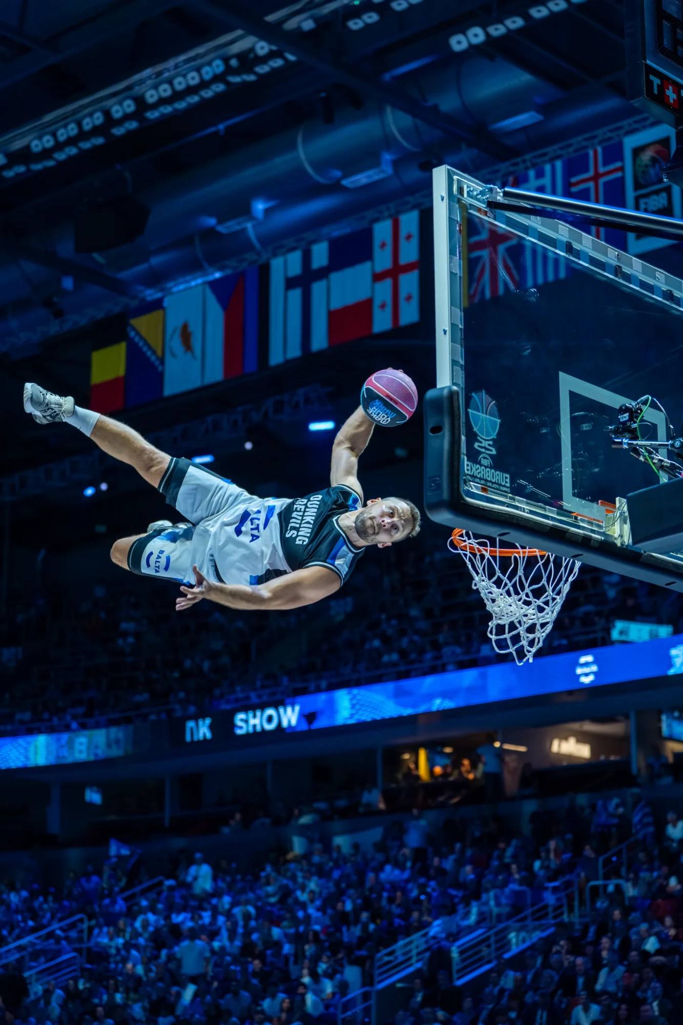 Ein Akrobat der Dunking Devils streckt sich während der Halbzeitshow in der Balta-Halle bei der EuroBasket 2025 neben dem Korb in der Luft.