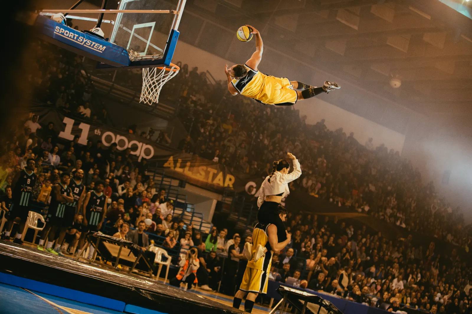 Ein Akrobat der Dunking Devils fliegt beim OPAP All-Star Game flach über das Spielfeld zum Dunk – vor voller Halle, die Spieler schauen von der Bank zu.