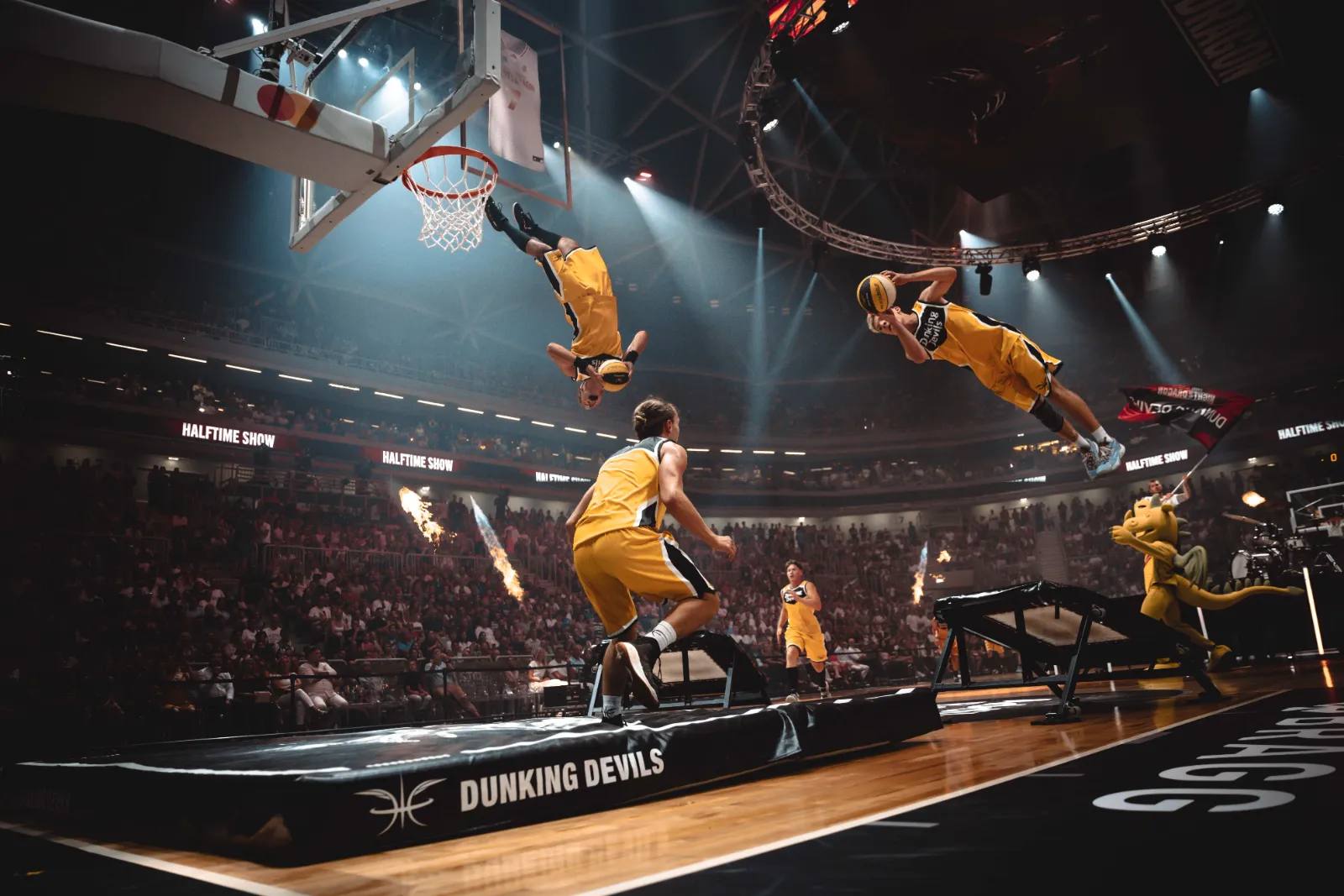 Zwei Akrobaten der Dunking Devils springen bei der Night of Dragon gleichzeitig mit Salto zum Dunk, auf dem Spielfeld das Team-Maskottchen und Flammen.