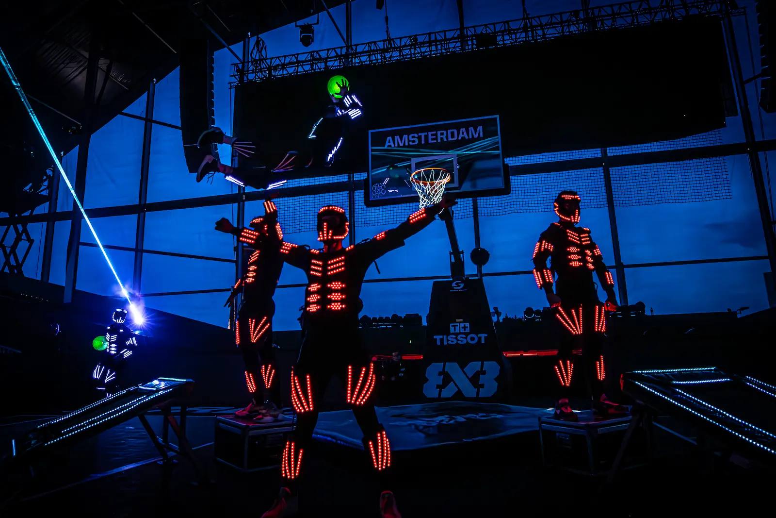 Dunking Devils LED acrobatic show at FIBA 3x3 Amsterdam: performers in glowing red LED suits on stage while one flies towards the hoop with a glowing ball.