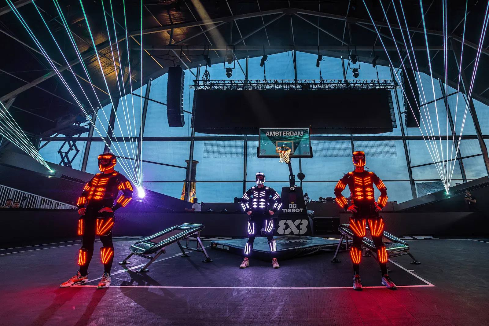 Three Dunking Devils performers in glowing LED suits posing under laser beams in front of the hoop.
