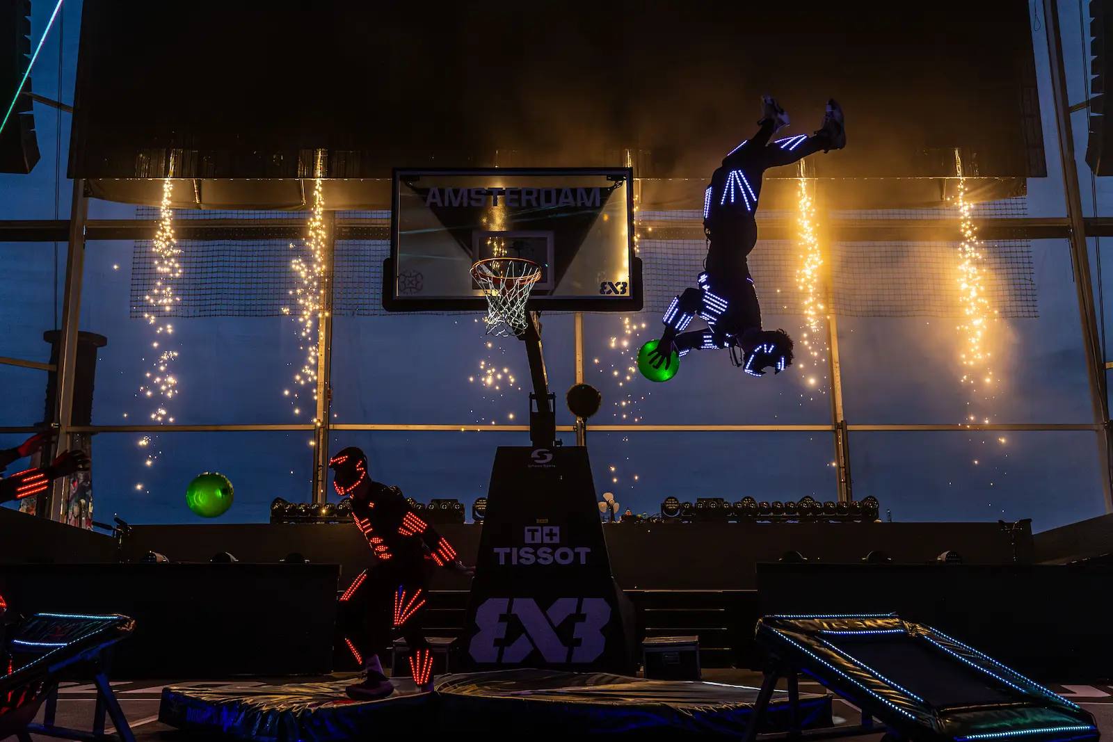 LED performer upside down mid-flip with a glowing green ball next to the hoop, with sparkler effects behind.