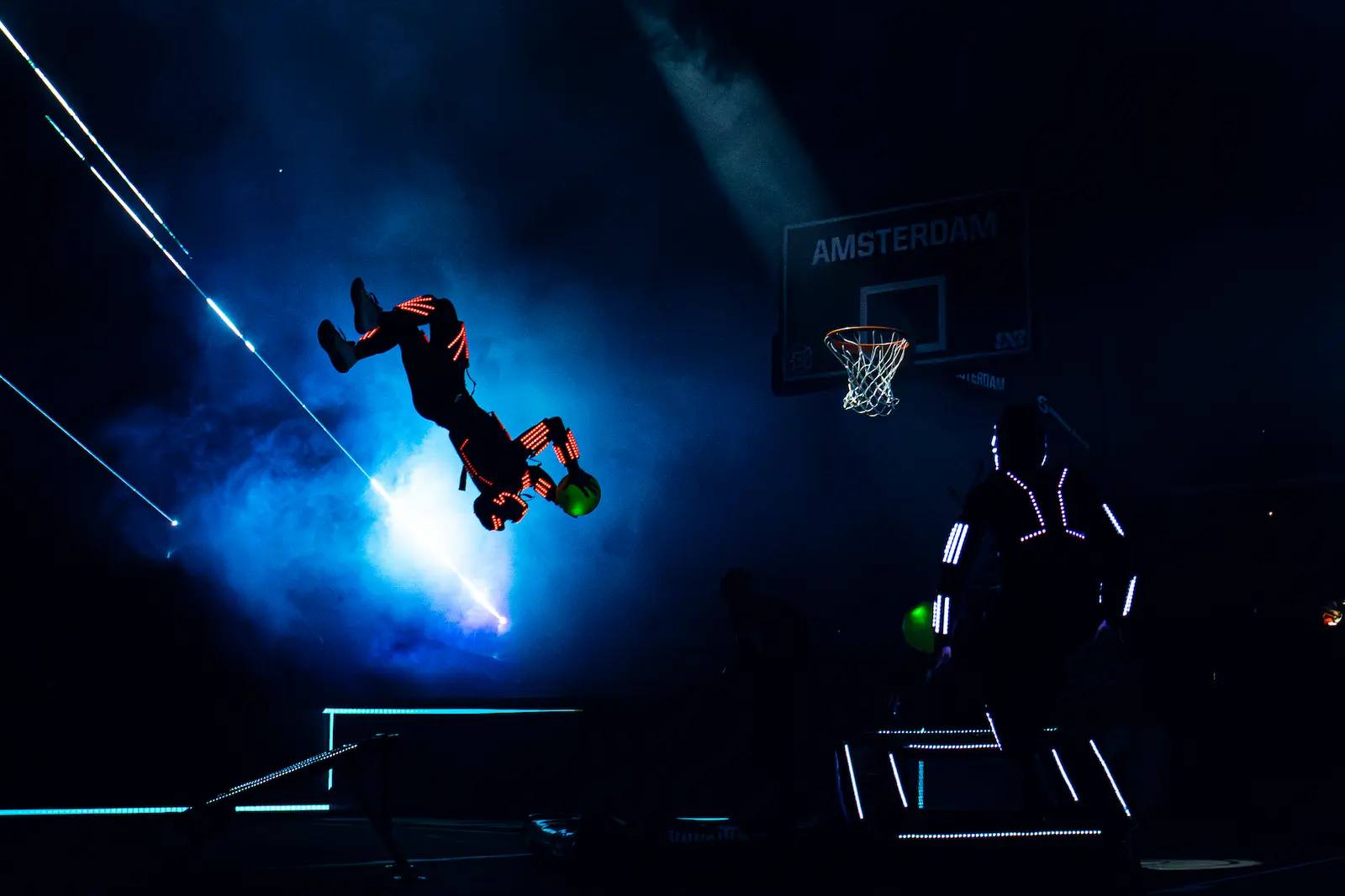 Red-lit LED acrobat flipping through blue haze and laser light towards the Amsterdam hoop.