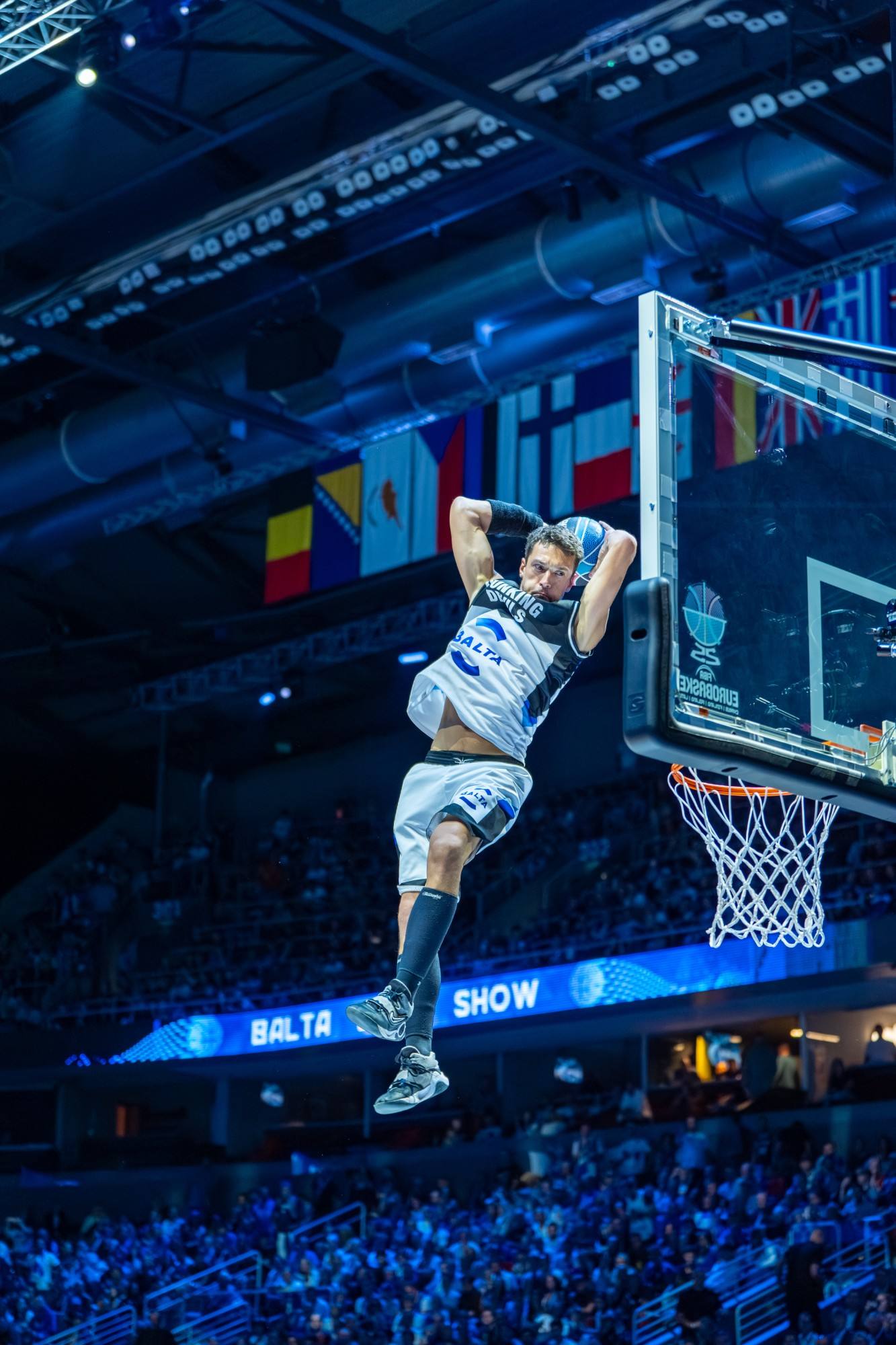Dunking Devils athlete hanging in the air before a dunk at EuroBasket 2025, under the arena lights and national flags.