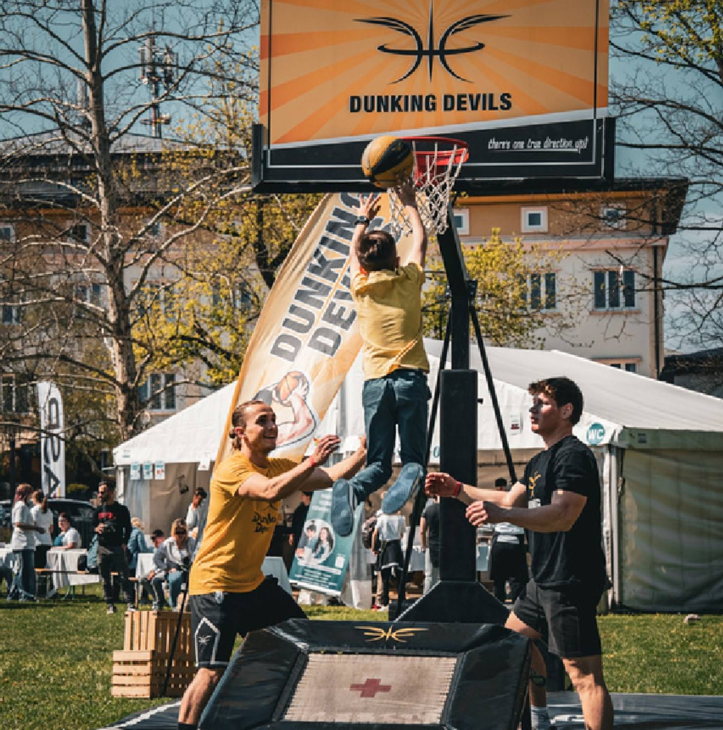 A young participant dunks off a mini trampoline at the Dunking Devils dunk station while coaches help.
