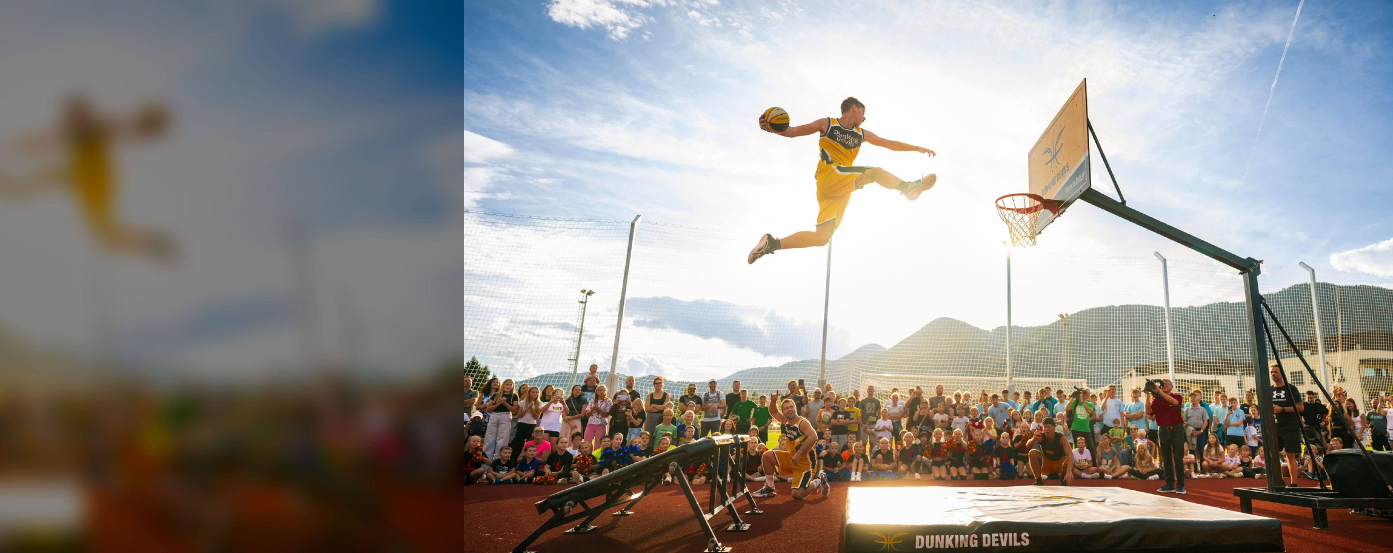 A Dunking Devils acrobat flies towards the hoop for a dunk at an outdoor show in front of a crowd of children.