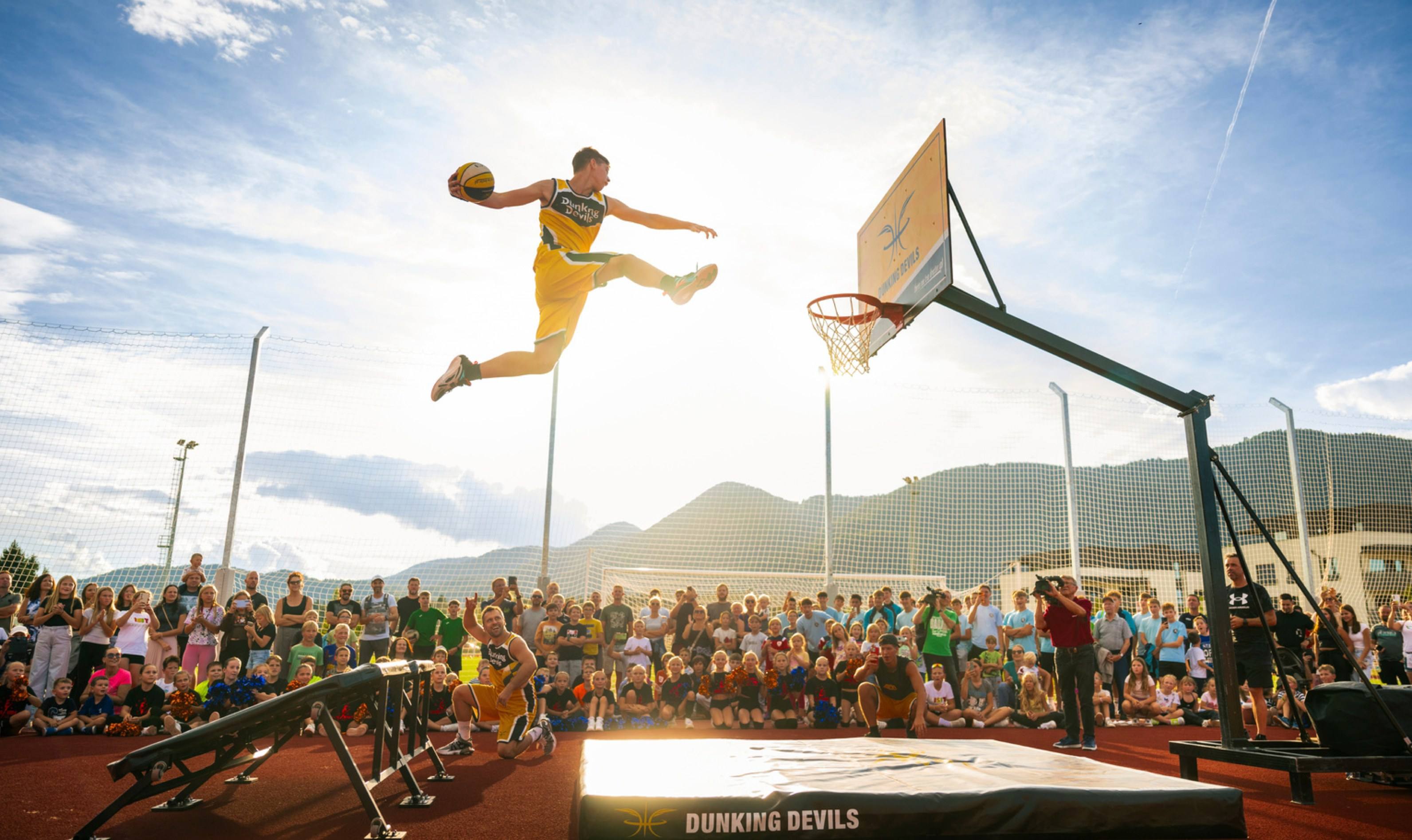 A Dunking Devils acrobat flies towards the hoop for a dunk at an outdoor show in front of a crowd of children.