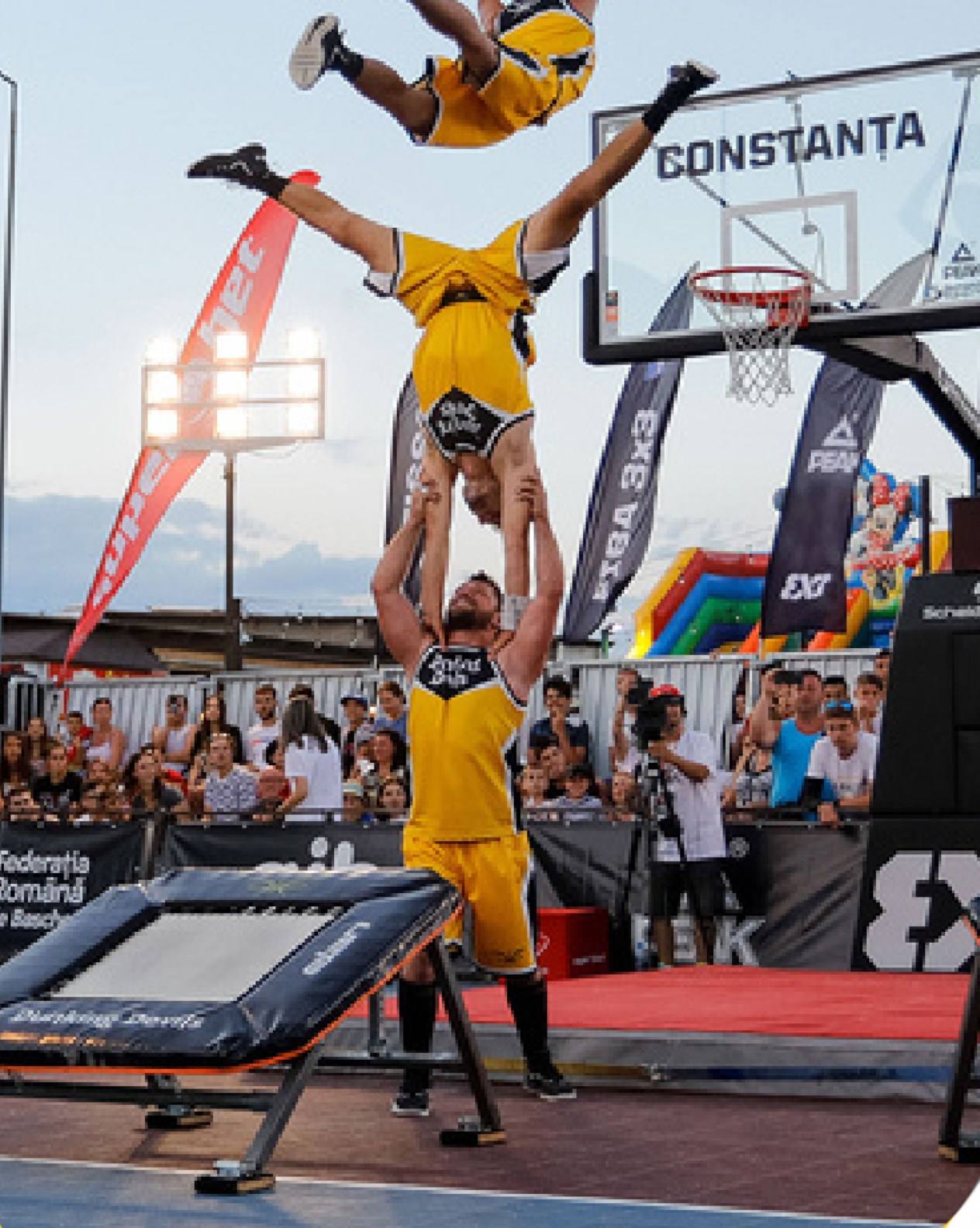 Dunking Devils acrobats build a human tower while a flyer flips above them at an outdoor 3x3 basketball event.