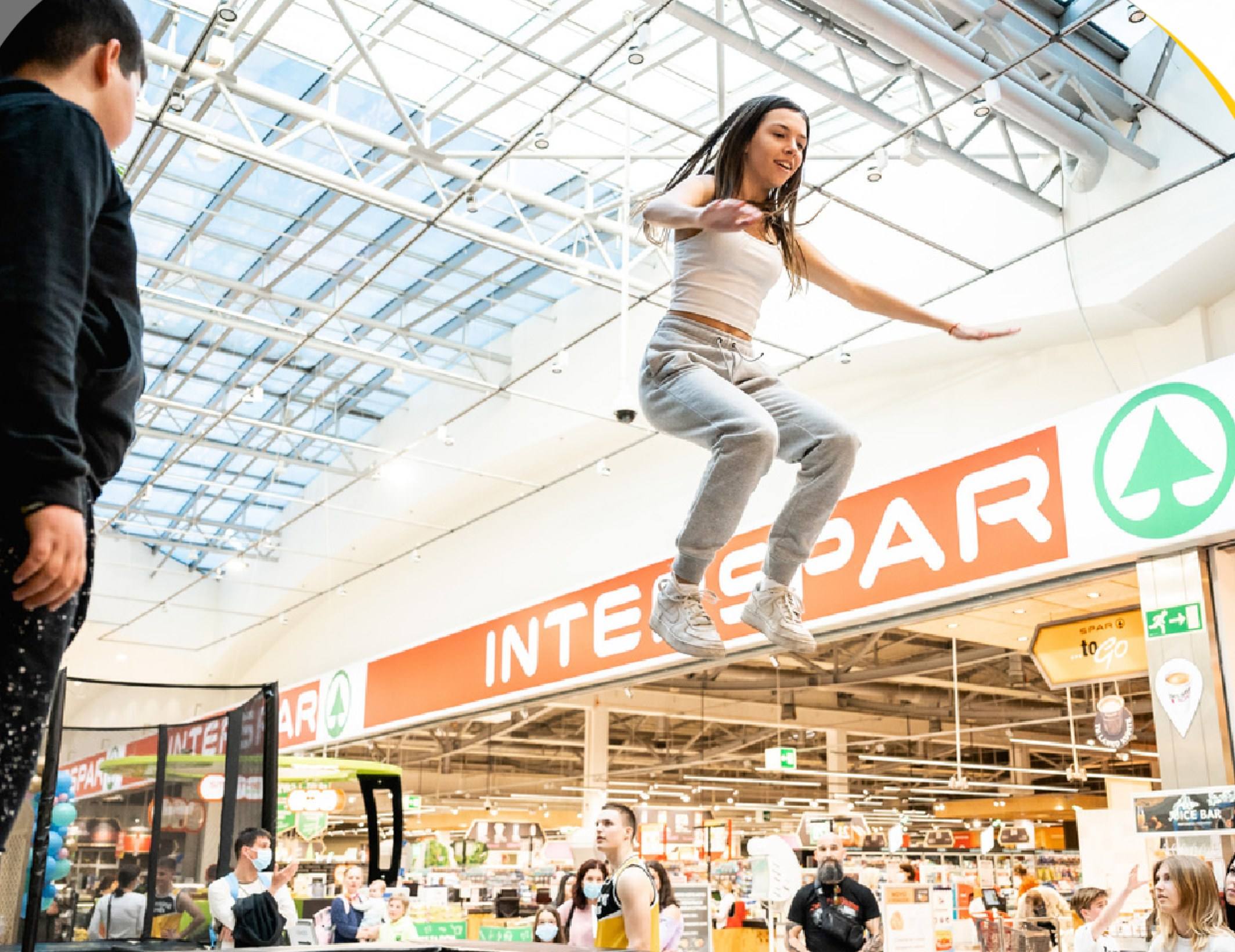 A girl jumps high on a trampoline under a glass roof at a shopping centre while people watch.