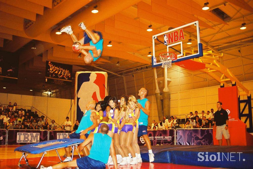 Archival photograph: a Žabe acrobat flips over a line of performers from a mini-trampoline towards an NBA-branded basket.
